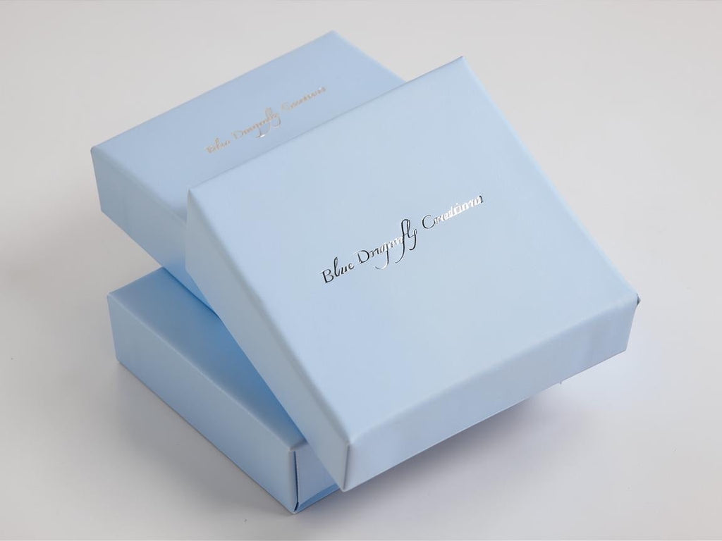 Blue Dragonfly Creations branded light blue gift boxes for jewelry packaging stacked on a white surface