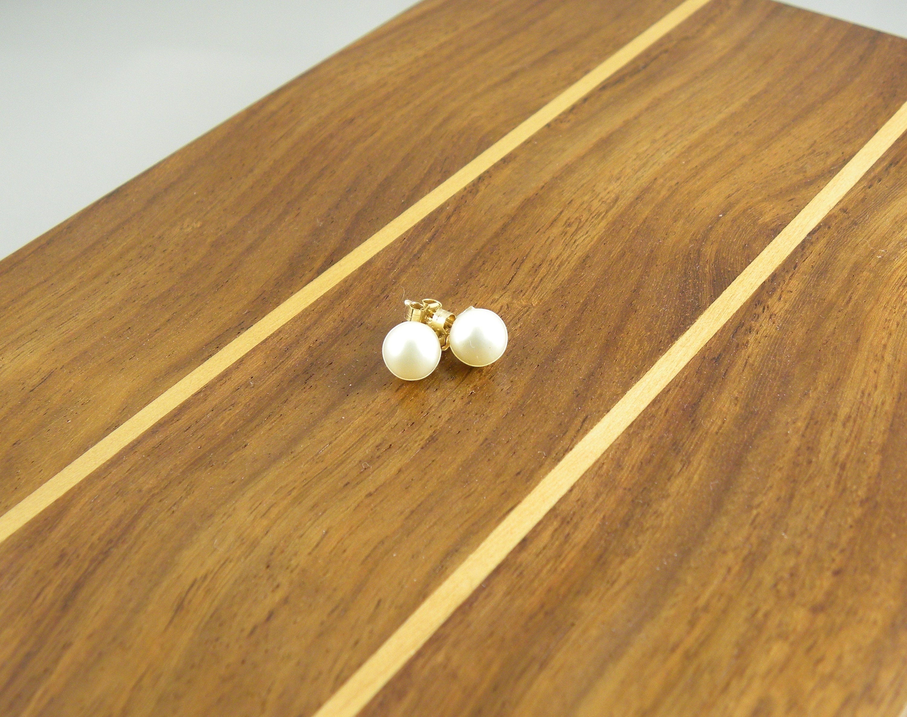 Blue Dragonfly Creations classic round pearl stud earrings with gold-tone posts resting on a polished wooden surface.